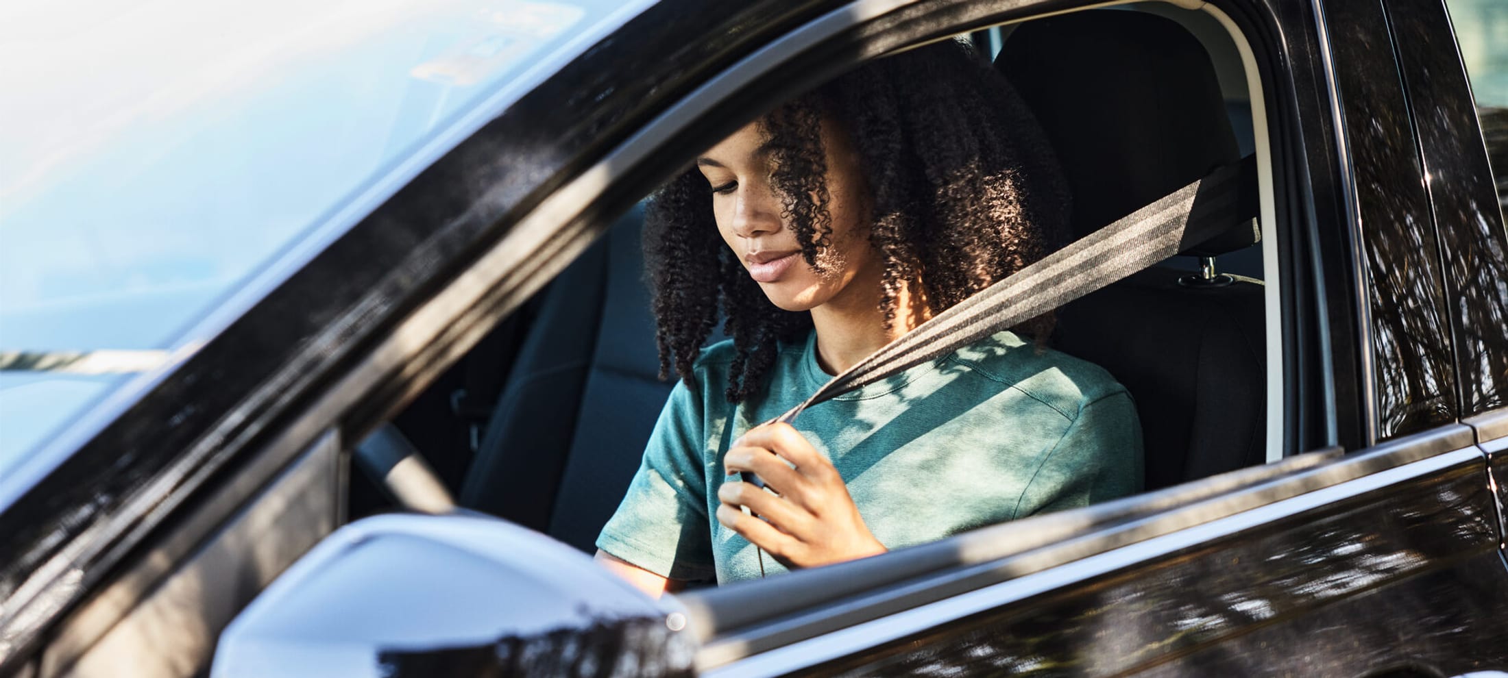 Una giovane donna siede al volante di un’auto nera e si allaccia le cinture di sicurezza. Il sole e un albero si riflettono nell’auto. 