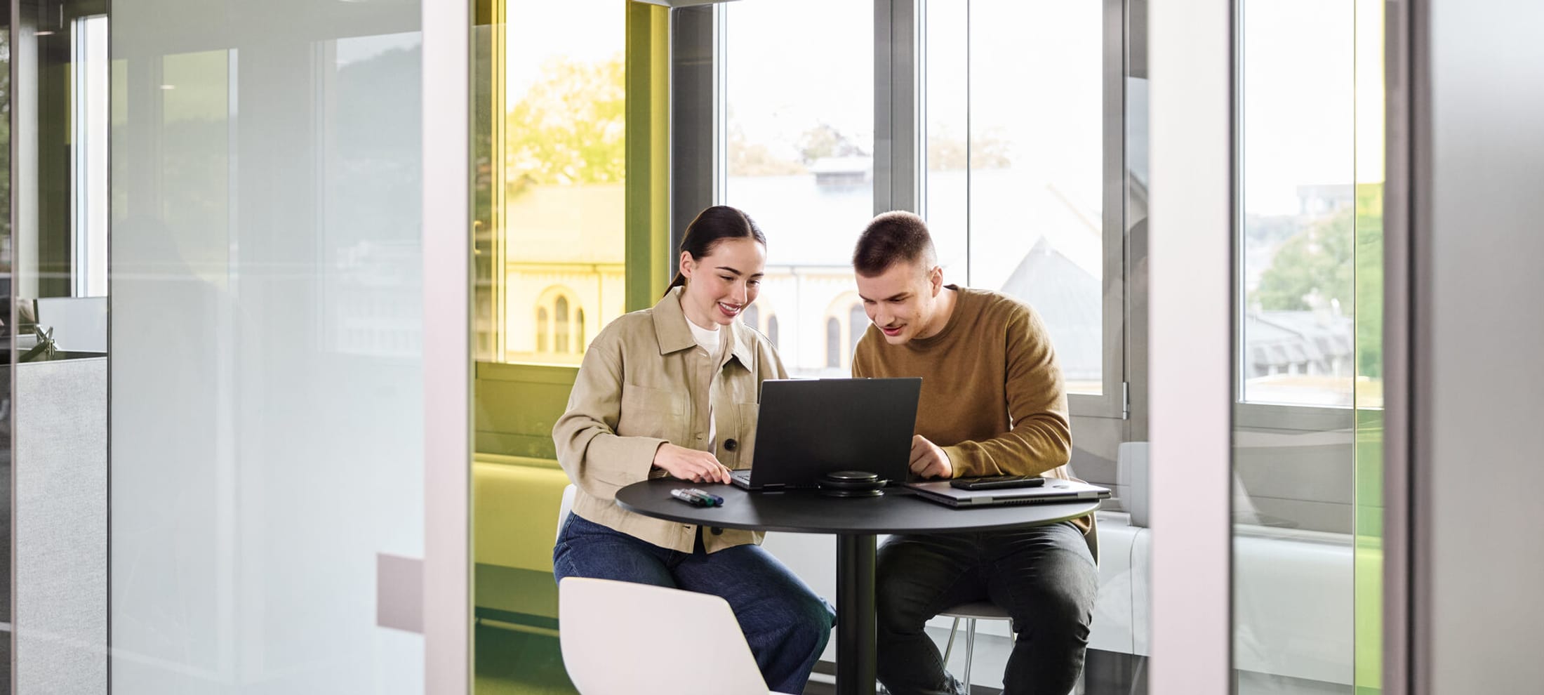 Un homme et une femme discutent debout, tandis qu’une autre femme travaille sur son ordinateur portable posé sur une table haute.