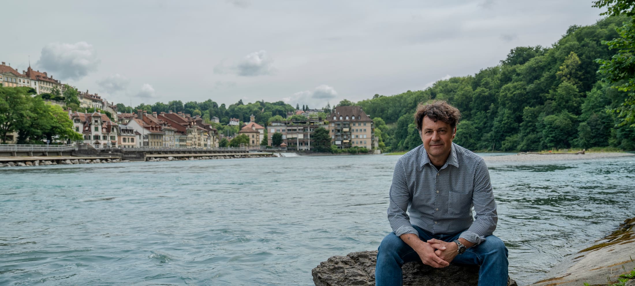 Andreas Zischg, Co-Leiter des Mobiliar Lab für Naturrisiken, sitzt auf einem Stein am Aareufer in Bern.