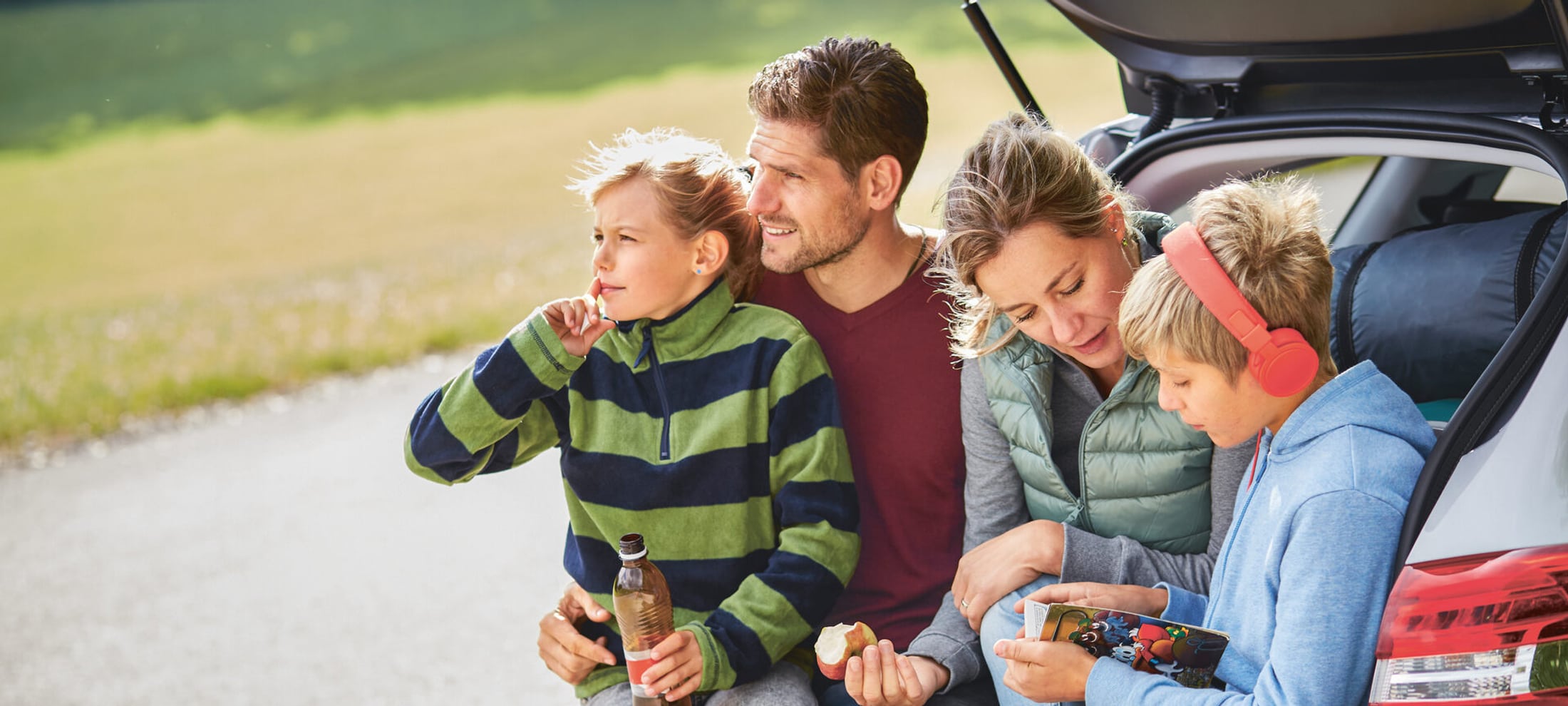 Familie mit Mutter, Vater, Tochter und Sohn sitzt bei offenen Kofferraumdeckel im Heck ihres Fahrzeugs.