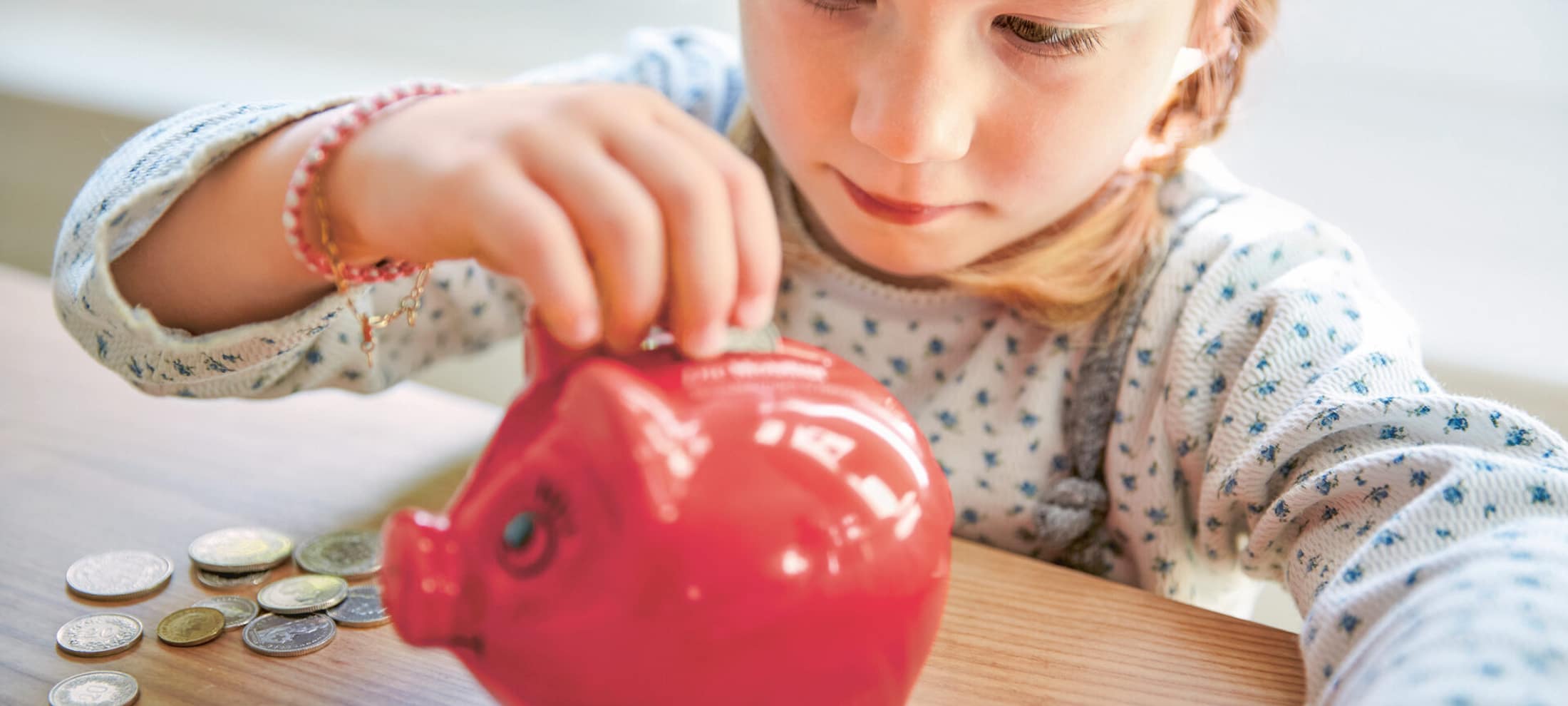 Una bambina siede a un tavolo su cui sono appoggiate alcune monete e ne infila una in un salvadanaio. 
