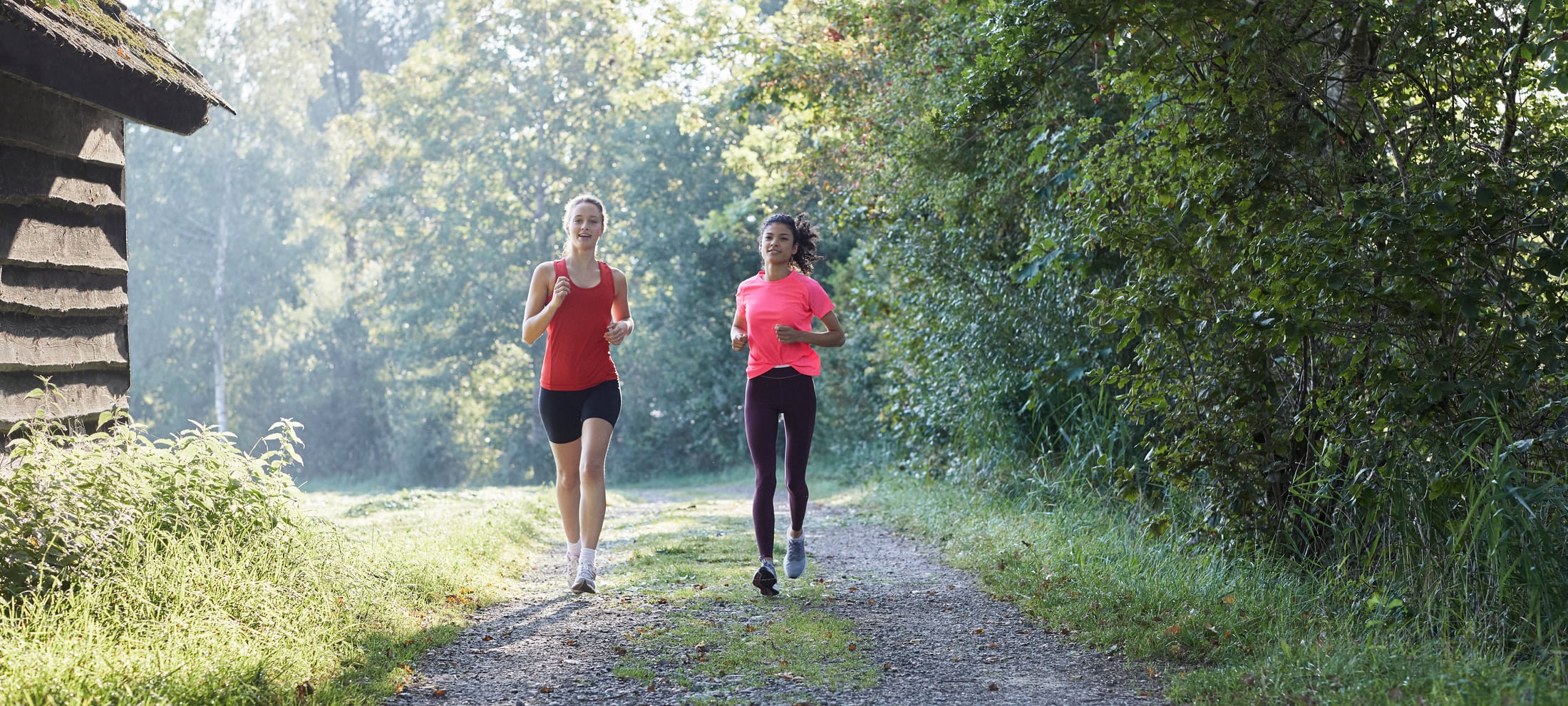 Zwei junge Frauen joggen bei Sonnenschein einem Waldrand entlang. 