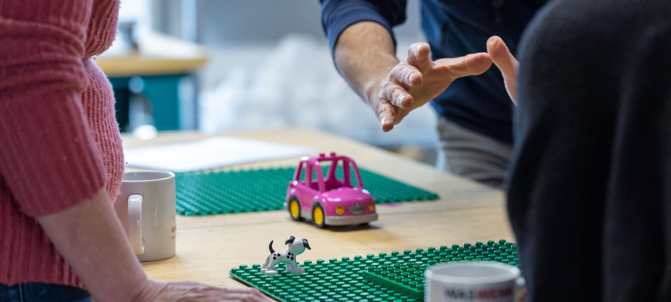 Foto dettagliata di una scrivania con base e personaggi della Lego, circondata dalle partecipanti e dai partecipanti al workshop. 