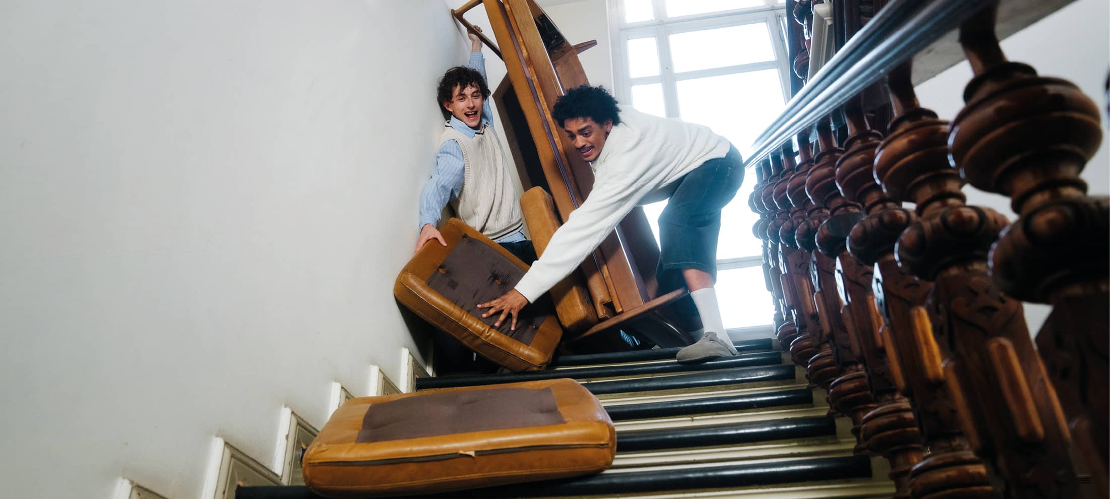 Zwei junge Männer manövrieren mit sichtlicher Mühe ein Sofa eine enge Treppe hinunter.