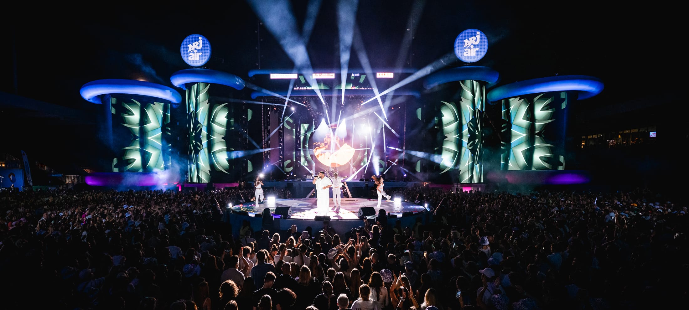 NRJ Air Bühne im Dunkeln mit Lichtshow und Sänger. Davor das Publikum von hinten.