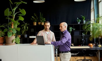 Due colleghi di lavoro parlano in piedi e guardano il laptop posizionato davanti a loro su un divisore. 