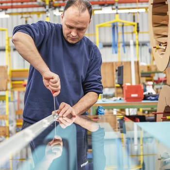Ein Mann in Arbeitskleidung arbeitet in einer Produktionshalle und schraubt etwas auf eine Glasplatte. 
