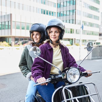 Zwei junge Frauen sitzen mit aufgesetztem Helm auf einem Motorroller und machen sich bereit zum Losfahren. 