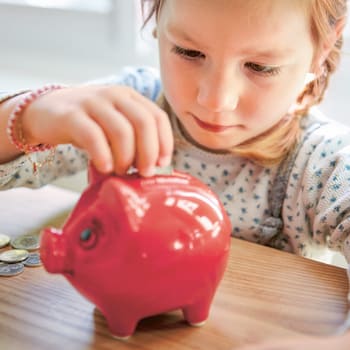 Ein kleines Mädchen sitzt an einem Tisch mit einigen Münzen und steckt eine von ihnen in ein Sparschwein. 