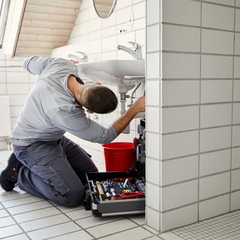 Ein Sanitärinstallateur repariert in einem Badezimmer kniend die Anschlüsse eines Lavabos. 