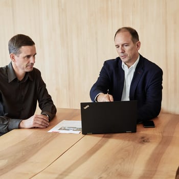 Due uomini sono seduti a un tavolo di legno in una sala riunioni e guardano insieme qualcosa sul laptop. Uno indica lo schermo del laptop. 