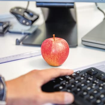 Dans le cadre de l’image, on voit un clavier posé sur un bureau, une main posée sur le clavier et une pomme rouge à côté du clavier. 