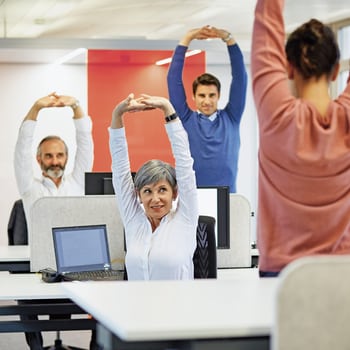 Dans un bureau, trois collaborateurs effectuent des exercices d’étirement sous la supervision d’une quatrième personne. 