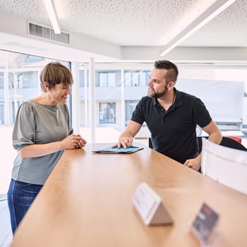 Ein Mann hinter einem Kundentresen gibt einer Frau eine blaue Arbeitsmappe. Beide lachen. 
