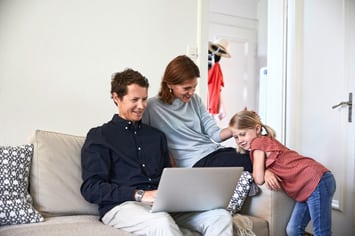 Ein Paar sitzt auf dem Sofa, ihre Tochter stützt sich darauf. Der Vater und die Tochter schauen auf einen Laptop, der auf den Oberschenkeln des Vaters liegt. Die Mutter lächelt ihre Tochter an und streicht ihr über den Kopf.
