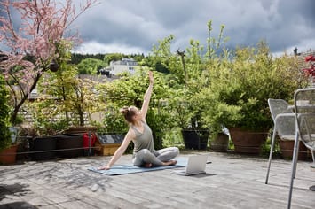 Eine junge Frau in Trainingskleidern sitzt in einer Yoga-Pose auf einer begrünten Terrasse auf einer Yoga-Matte. Vor ihr liegt ein aufgeklappter Laptop am Boden. 