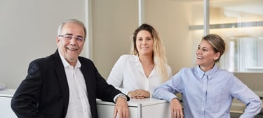 Un homme âgé et deux jeunes femmes se tiennent debout autour d’un cube de bureau. Ils rient et s’appuient les coudes contre le cube. 