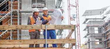 Zwei Männer mit Bauhelmen stehen auf einer Baustelle und sehen sich gemeinsam die Baupläne an. 