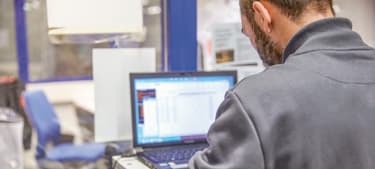 Ein Mann steht vor einem Laptop und schreibt etwas auf, neben ihm liegt ein Anzeige- oder Diagnosegerät. 