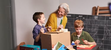 Une femme âgée et ses deux petits-enfants se tiennent devant une ardoise dans une salle de bricolage, à une table entourée d’un carton et d’autres ustensiles de bricolage.