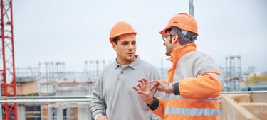 Zwei Männer stehen auf einer Grossbaustelle und sprechen zusammen. Sie tragen beide Schutzhelme und ein Mann trägt eine Sicherheitsweste. 