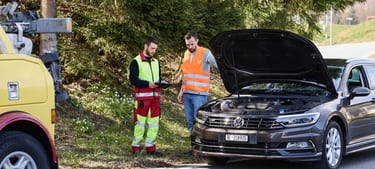 Zwei Männer in Warnkleidung stehen neben einem Auto mit offener Motorhaube und unterhalten sich.