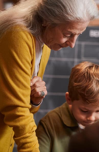 Una donna anziana è seduta ad un tavolo da bricolage con il nipote e lo guarda da dietro le spalle.