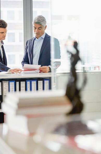 Due uomini in giacca e cravatta si consultano su questioni legali, in primo piano si vede sfocata una figura della giustizia. 