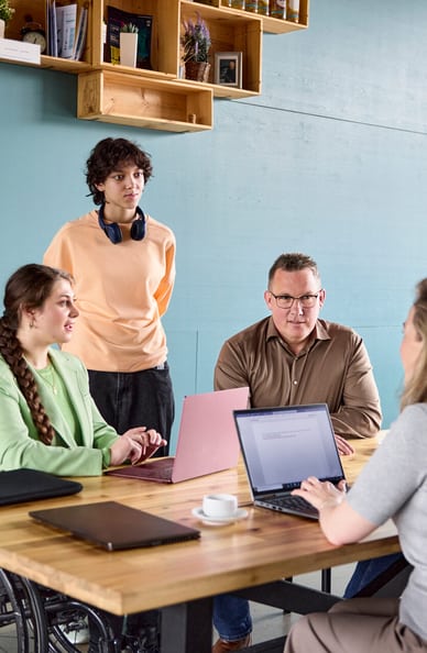 A un tavolo di legno siedono quattro persone vis-à-vis, una donna in sedia a rotelle. Un’altra donna è dietro i due partecipanti un po’ più distanti. Due persone al tavolo hanno un laptop aperto davanti a sé e scrivono.  