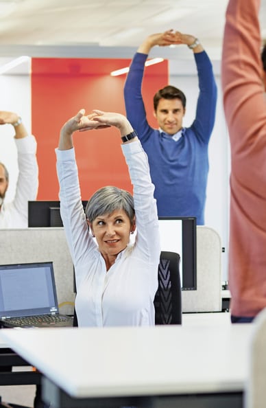 In un ufficio tre collaboratori fanno esercizi di stretching sotto la guida di una quarta persona. 