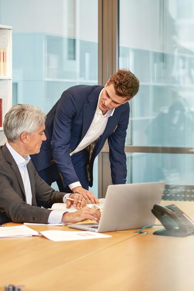 Due uomini sono in un ufficio, uno seduto al tavolo delle conferenze e l’altro accanto. Sul tavolo ci sono delle foglie e un portatile. I due uomini stanno discutendo.