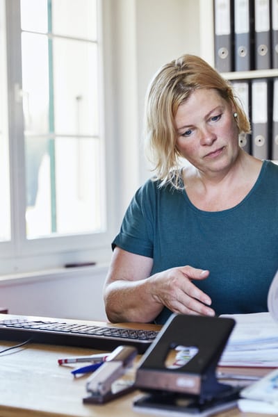 C’e’ una donna seduta alla sua scrivania in ufficio che sfoglia un raccoglitore federale per cercare qualcosa. In primo piano si vedono il suo computer e piccoli utensili da ufficio.