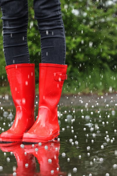 Si vede solo la parte inferiore delle gambe di un adulto che indossa stivali di gomma rossi, i piedi sono in una pozzanghera e piove la grandine. 