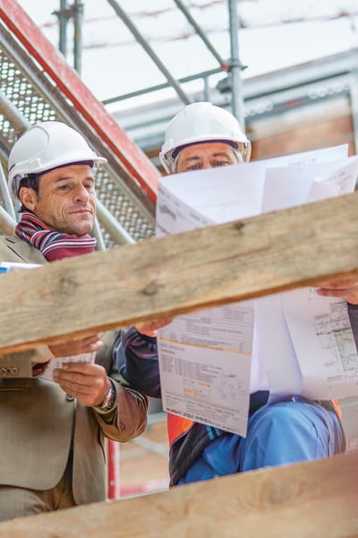 Zwei Männer stehen auf dem Gerüst einer Baustelle und schauen auf einen Bauplan.