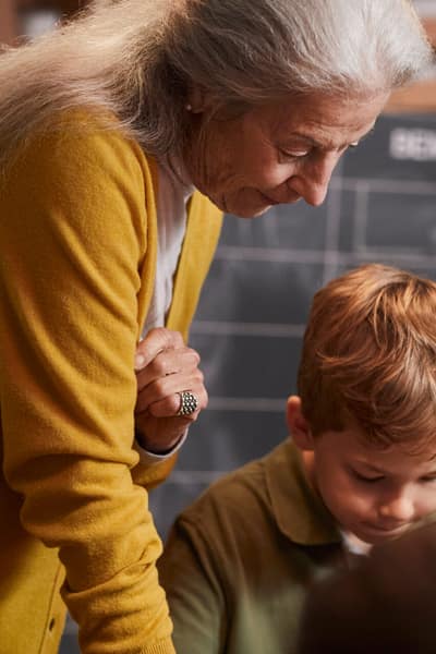 Une femme âgée se tient à une table de bricolage avec son petit-fils et regarde par-dessus son épaule.
