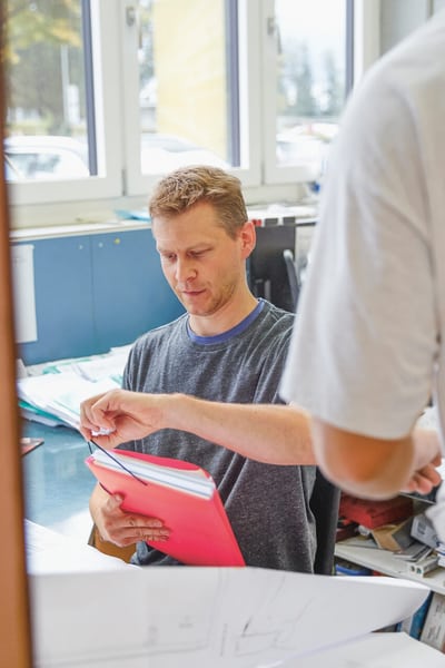 Ein Mann sitzt in einem Büro in einer Schreinerei und sucht unterlagen heraus. Ein zweiter Mann spricht mit ihm.
