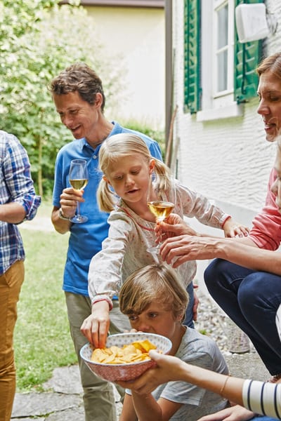 Zwei Männer, eine Frau, ein Teenager-Mädchen, ein Mädchen und ein Junge sind im Garten neben einem Grill versammelt. Sie lachen und reden. Die Erwachsenen trinken Wein, Jüngeren essen Chips aus einer Schüssel.