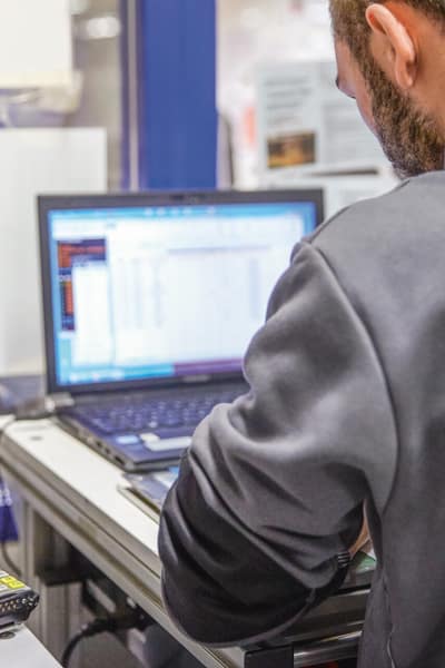 Ein Mann steht vor einem Laptop und schreibt etwas auf, neben ihm liegt ein Anzeige- oder Diagnosegerät. 