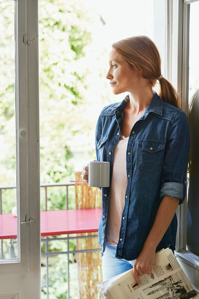 Une jeune femme se tient debout dans le cadre de la porte de son balcon, une tasse à café à la main et regarde au loin.