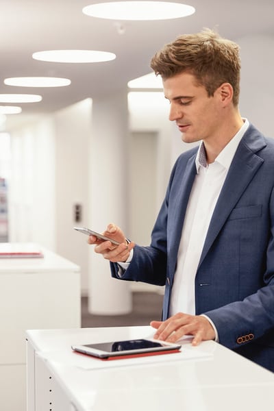 Ein Mann in Anzug blickt auf sein Handy. Er steht an einem Tresen in einem Büro.