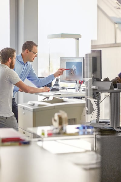 Zwei Männer stehen in einem Büro vor einem Bildschirm und besprechen etwas.