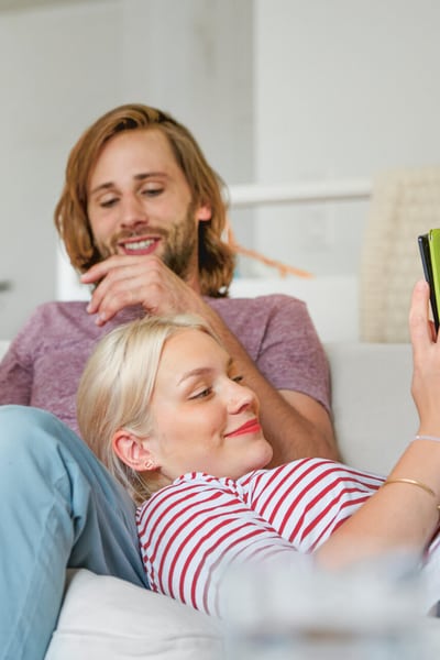 Ein Paar liegt auf dem Sofa in einem Wohnzimmer. Der Mann sitzt und die Frau liegt, ihren Kopf auf das Bein ihres Partners gelegt. Beide haben ein Handy in der Hand.