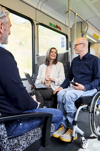 Eine Frau und zwei Männer sitzen in einem Zugabteil, lachen und reden zusammen. Die Frau hat ein Laptop auf dem Schoss, einer der Männer ein Smartphone in der Hand. 