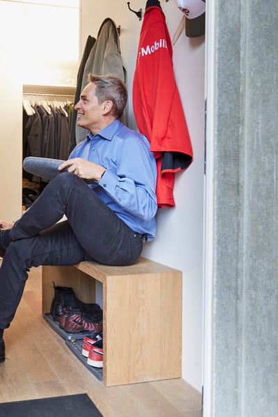 Mann sitzt in Garderobe und zieht sich Mobiliar Hausschuhe an. An der Garderobe hängt eine Mobiliar Jacke.