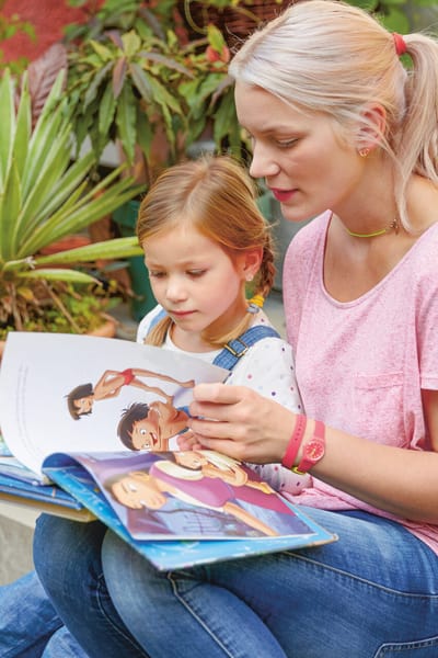 Une jeune femme est assise avec sa fille dans le jardin sur les escaliers menant à une porte d’entrée. Ils regardent ensemble un livre d’images. 