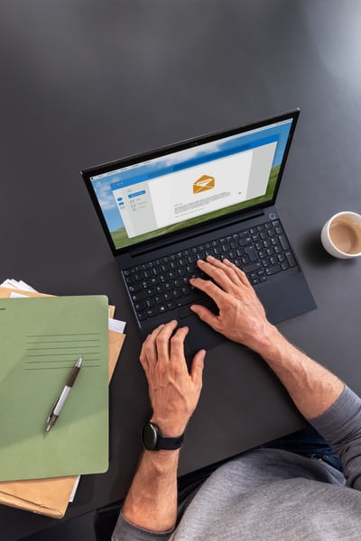 Person bedient einen Laptop auf dem Tisch. Daneben liegen ein Stapel Papier und eine Tasse Kaffee. 