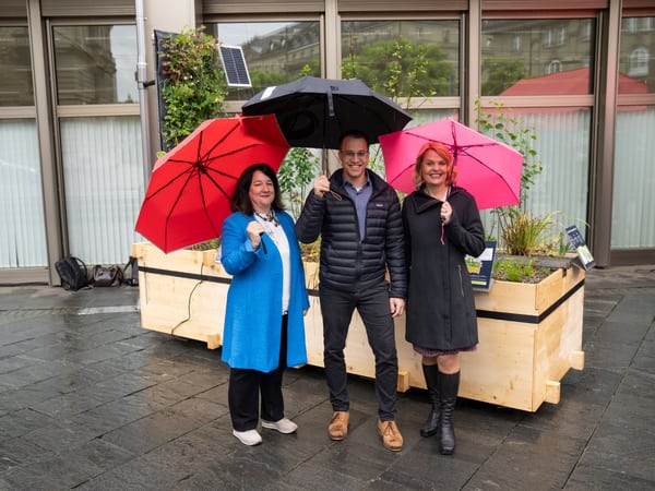 3 Personen mit einem Regenschirm in der Hand stehen vor dem Pflanzenbeet.