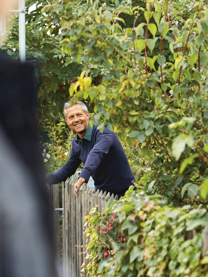 Ein Mann lehnt sich über einen Holzzaun, um ihn herum sind dichte grüne Pflanzen. Symbolbild für das Thema Einfriedung.