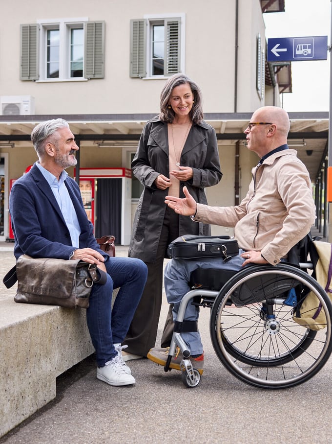 Ältere Freunde, darunter ein Mann im Rollstuhl, treffen sich am Bahnhof und sprechen miteinander