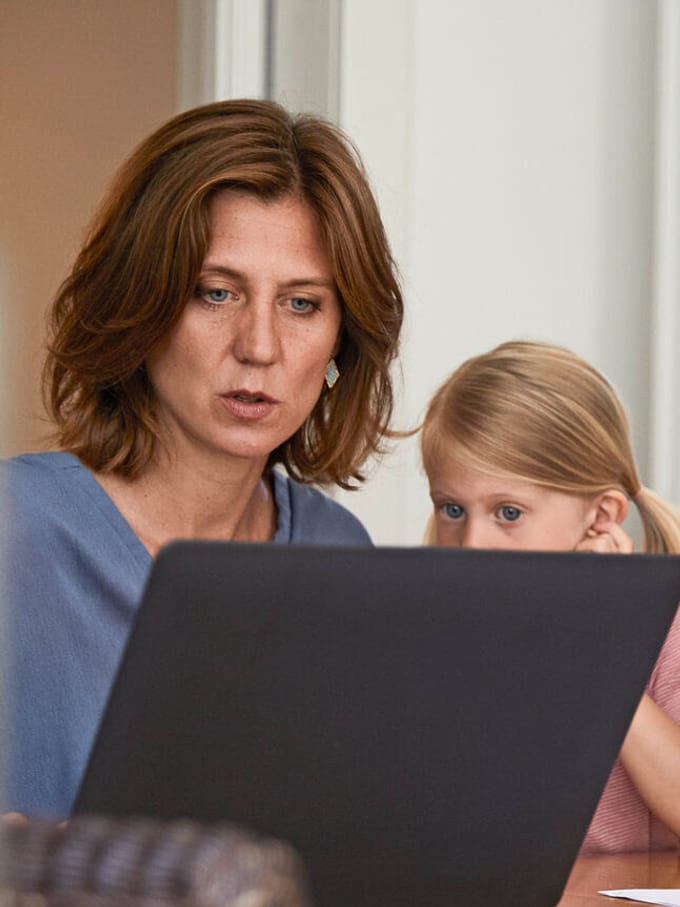 Frau sitzt an Laptop, etwas schein nicht zu stimmen. Kinder daneben eines an den Hausaufgaben, das andere schaut mit auf den Bildschirm.